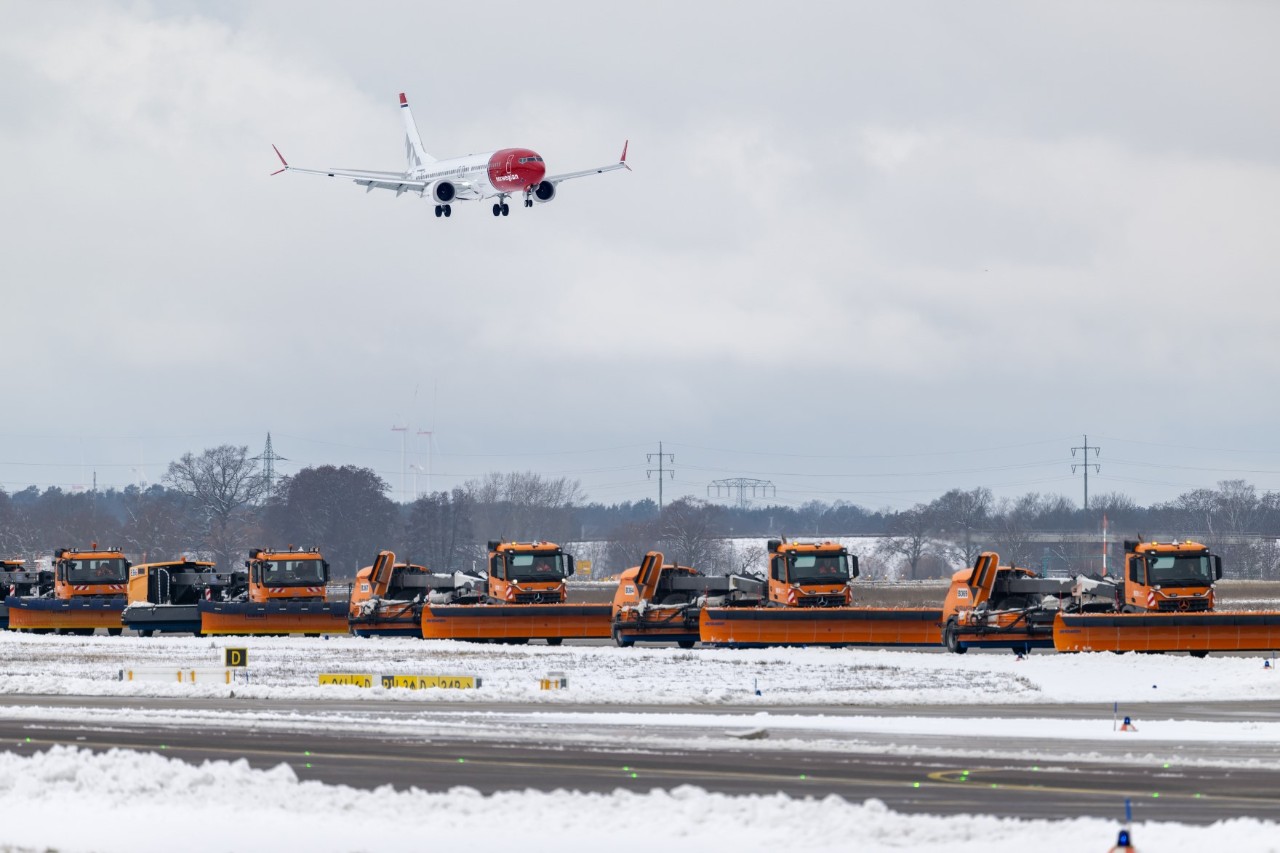 Ein Flugzeug der Airline Norwegian Airways im Landeanflug. Darunter viele Räumungsfahrzeuge, die in einer Reihe hintereinander fahren.