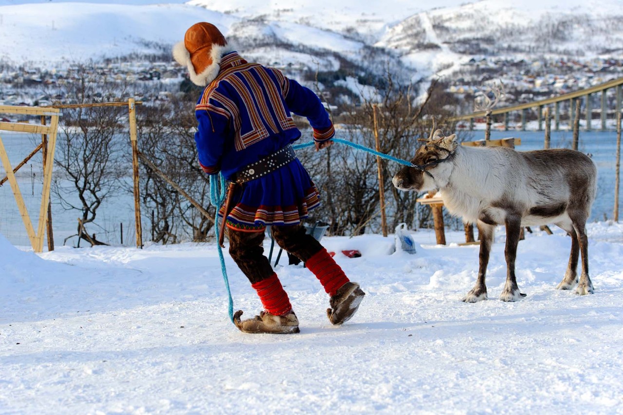 Ein Mann führt ein Rentier durch die verschneite Landschaft. Im Hintergrund erheben sich mit schneebedeckte Berge.