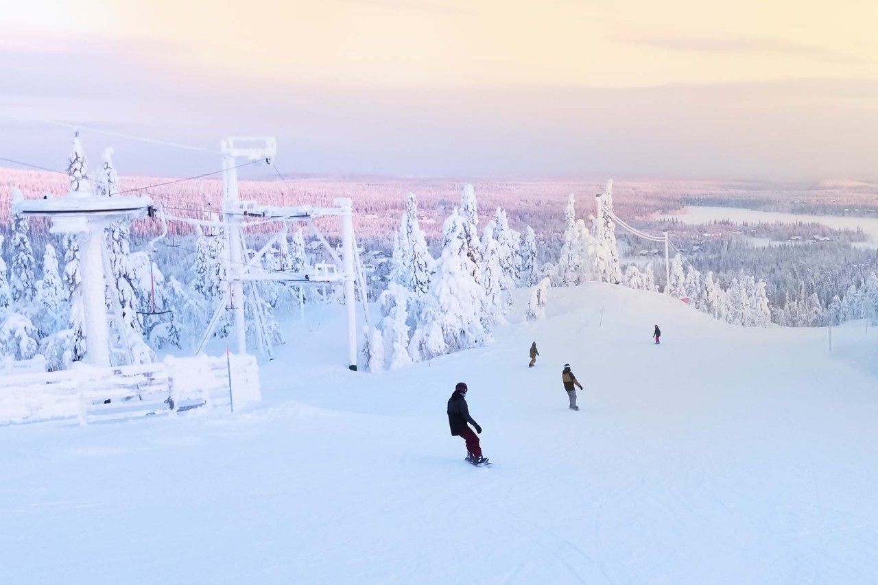 Eine Gruppe von Skifahrern und Snowboardern fährt den Berg hinab. Im Hintergrund geht die Sonne über einem schneebedeckten Wald auf. Panoramaansicht von Kuusamo–Ruka in Finnland.