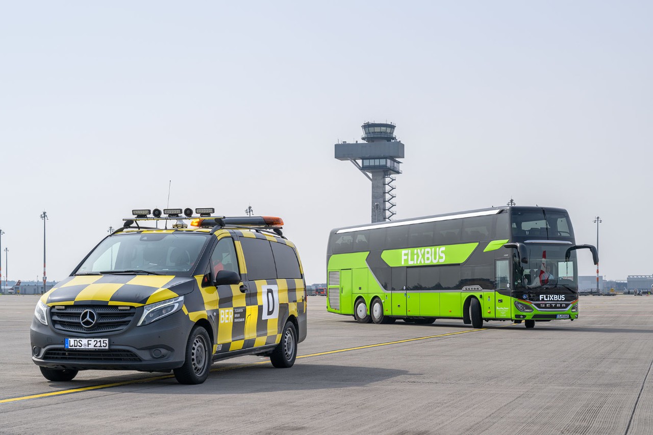 Ein Follow-Me-Auto steht auf dem Vorfeld. Im Hintergrund fährt ein grüner Fernbus. 