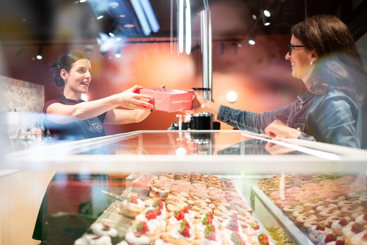 Eine Verkäuferin übergibt, über eine Theke voll mit Donuts hinweg, einer Kundin eine Box.