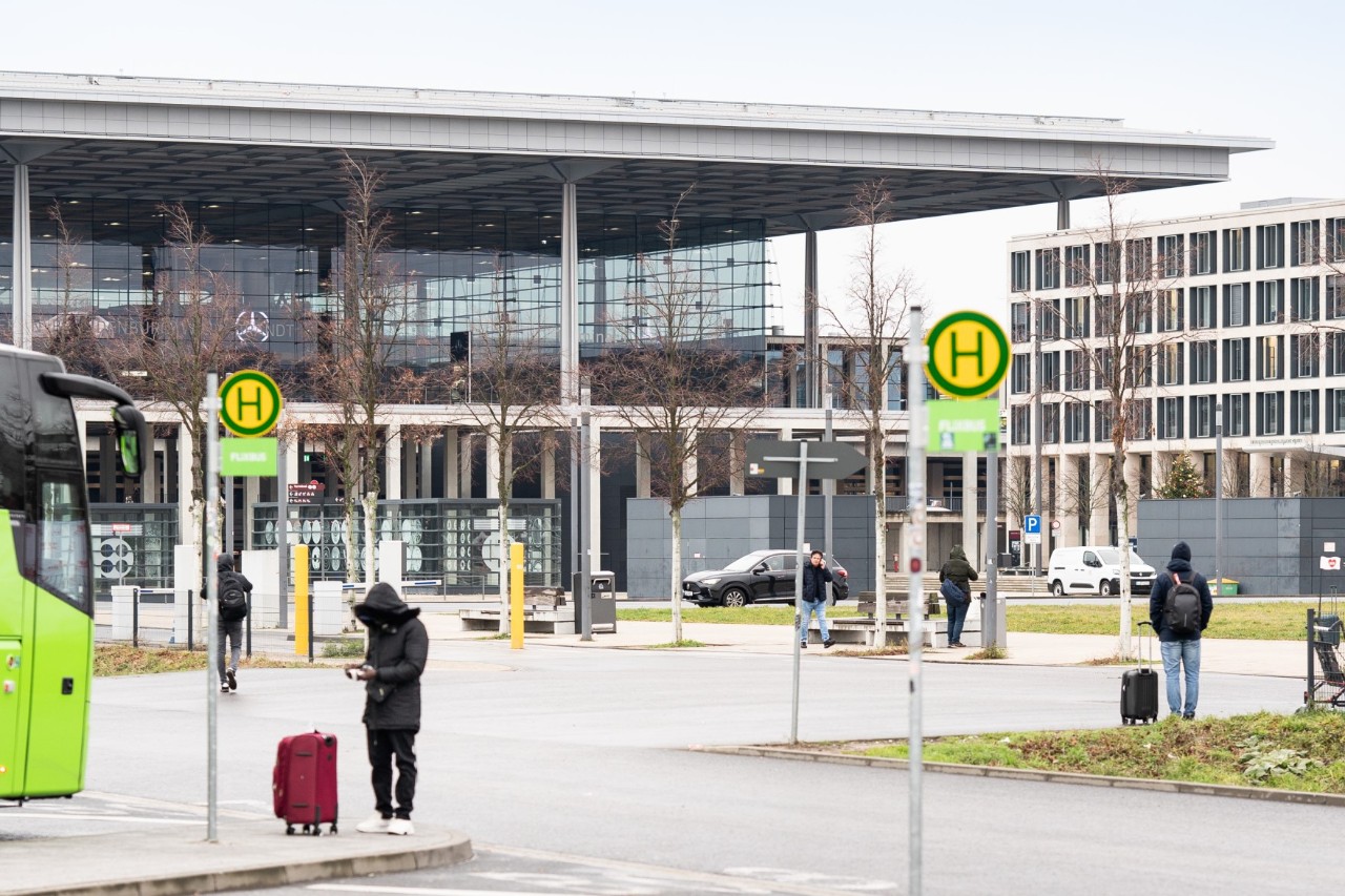Grüner Bus an der Bushaltestelle B. Daneben wartetende Personen und das Terminal 1.
