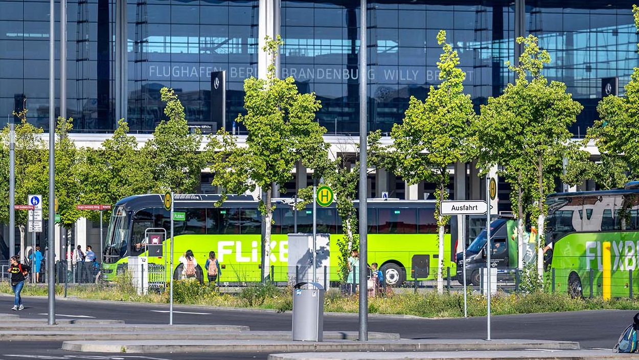 Grüner Bus an der Bushaltestelle B. Daneben wartetende Personen und das Terminal 1.