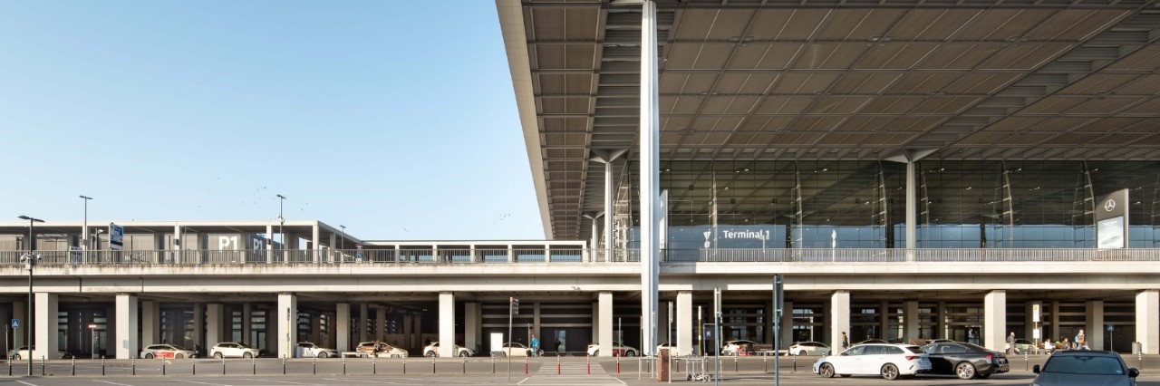 Frau steht mit Koffer und Kaffee in der Hand vor einem grauen Auto. Dahinter ist das Flughafen Terminal zu sehen.