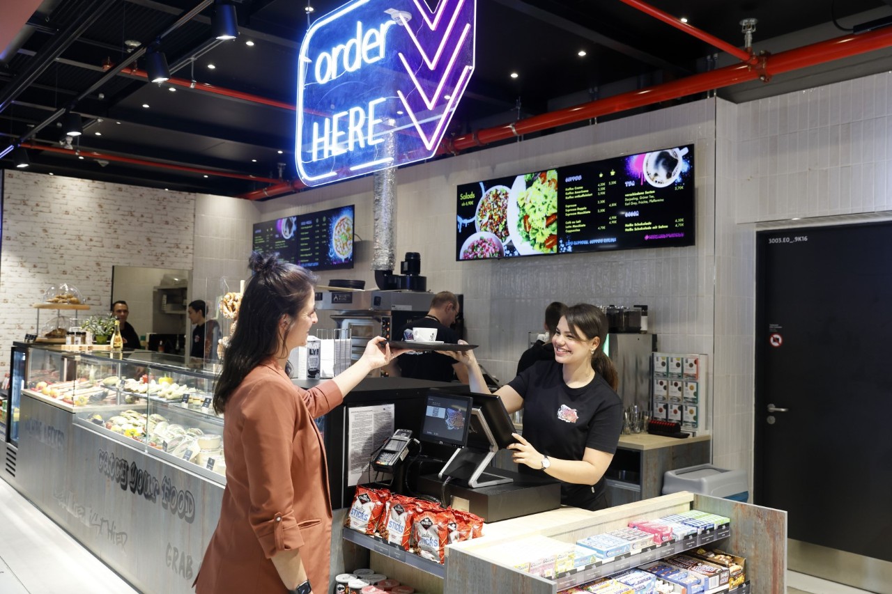 Counter of a catering establishment. A woman buys a coffee, which is handed to her by the cashier.