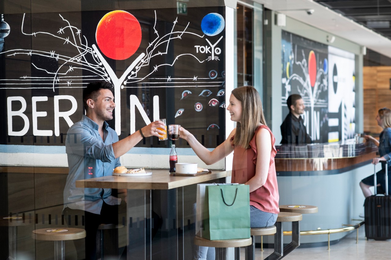 Two people are eating and drinking in a restaurant. Two people are talking in the background.
