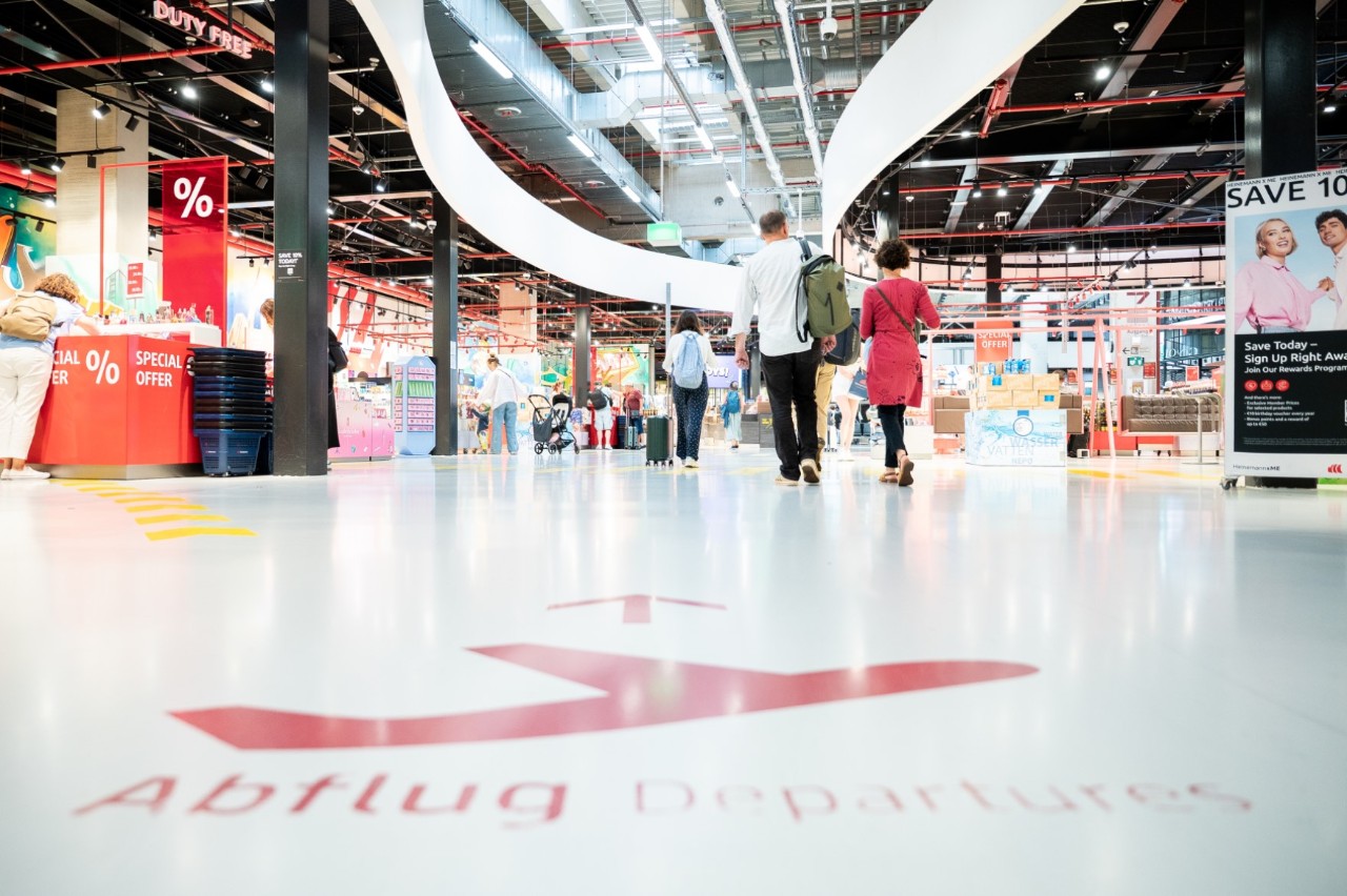People walk down an open corridor with signs pointing to the departure area. There are shelves with goods on the right and left.