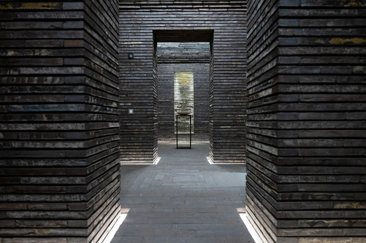 Interior shot of the Still room. Light shines into the room above. There is a candle at the back. The walls are made of brownish stone.