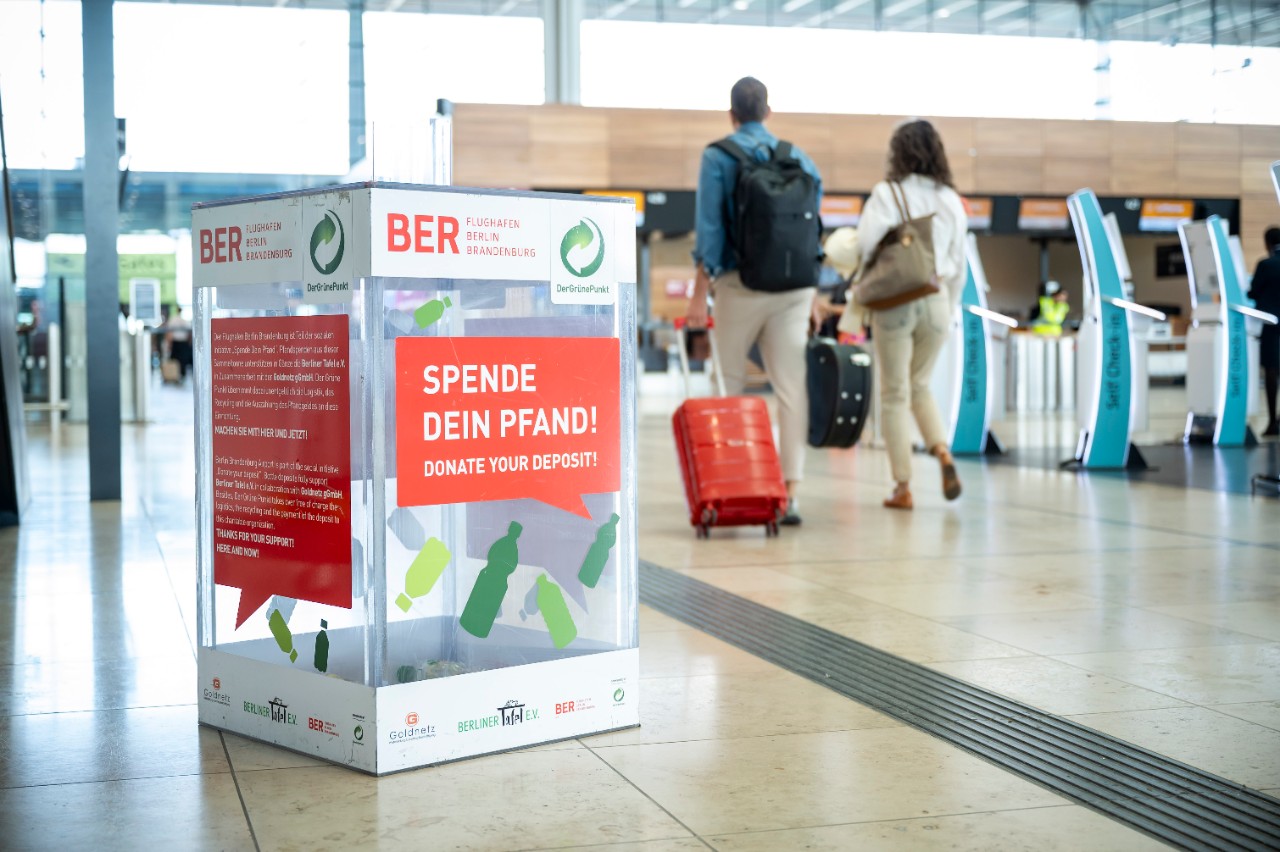 Donate your deposit at the bin in the check-in hall of BER. In the background, passengers with suitcases.