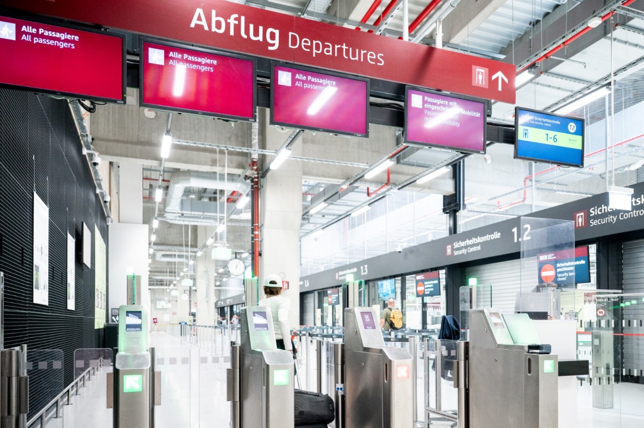 Security screening lanes in Terminal 2 of BER Airport, with monitors above and a glass door in the background.