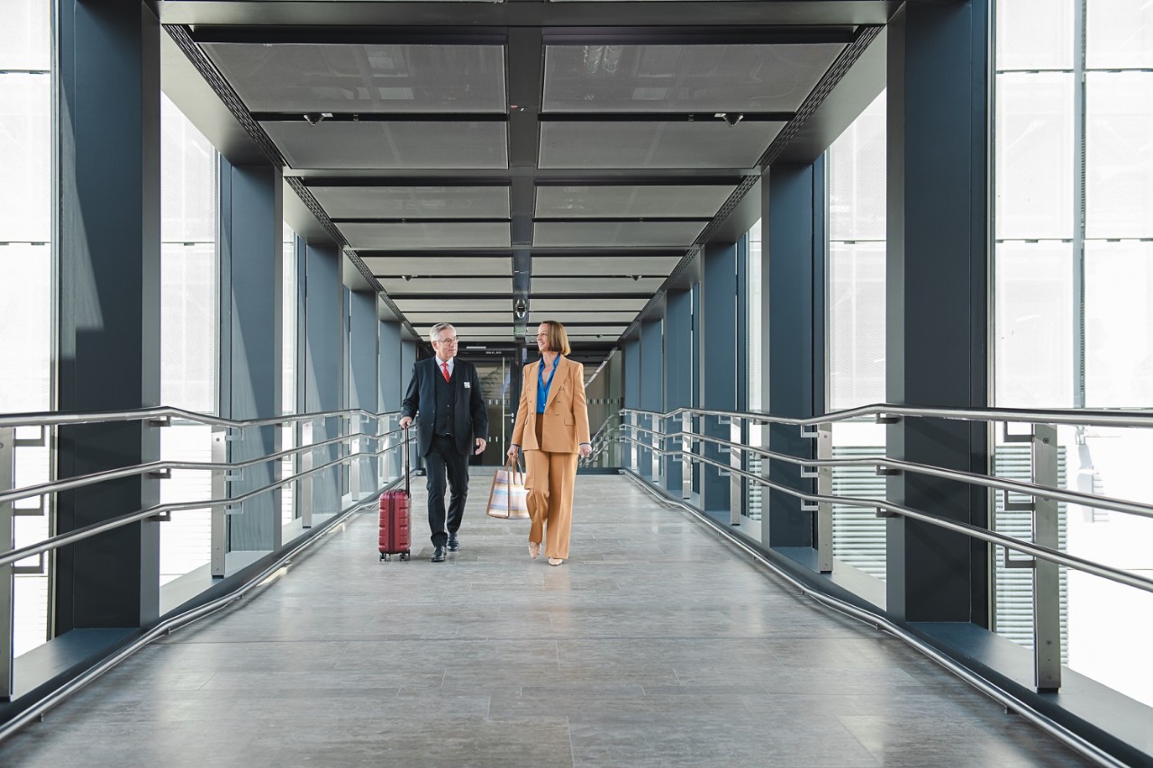 2 Personen gehen in einer Glasbrücke vom Parkhaus zur Lounge. Person 1 hat einen Koffer. Person 2 eine Tasche in der Hand.©Ekaterina Zershchikova / Flughafen Berlin Brandenburg GmbH