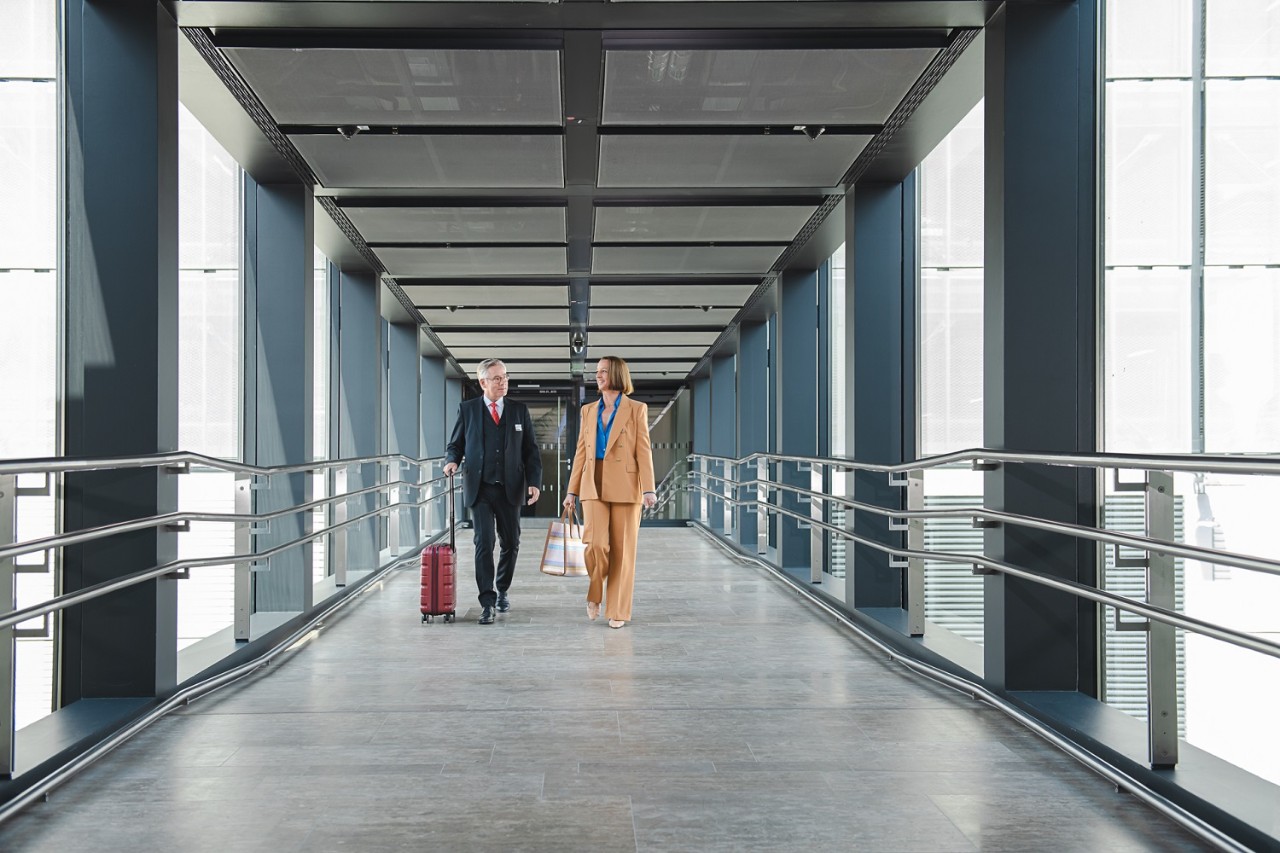2 people walk in a glass bridge from the car park to the lounge. 