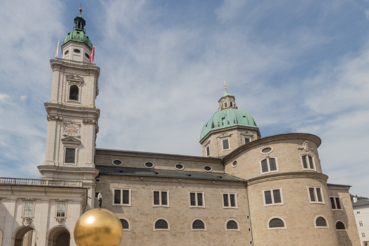 View of a baroque, sand-coloured building with towers and green domes. The sky is streaked with clouds. In front of the building on the left is a huge golden sphere with a very small person standing on it.