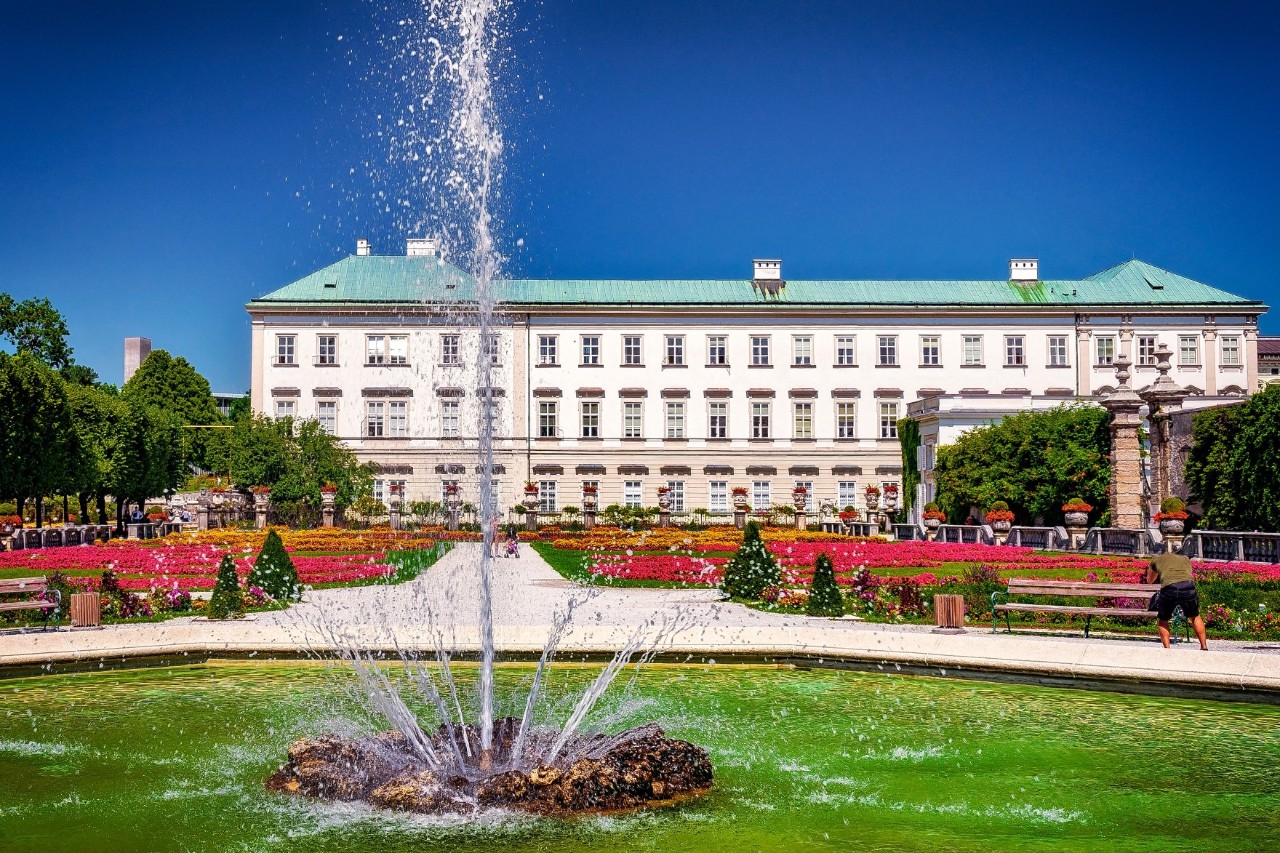 Frontal view of a three-storey, light-coloured palace with a light green roof, in front of it, to the left and right, red and yellow flowerbeds, in the middle a path leading to the palace. In the foreground is a water basin with a fountain. The sky is blue on a sunny day.