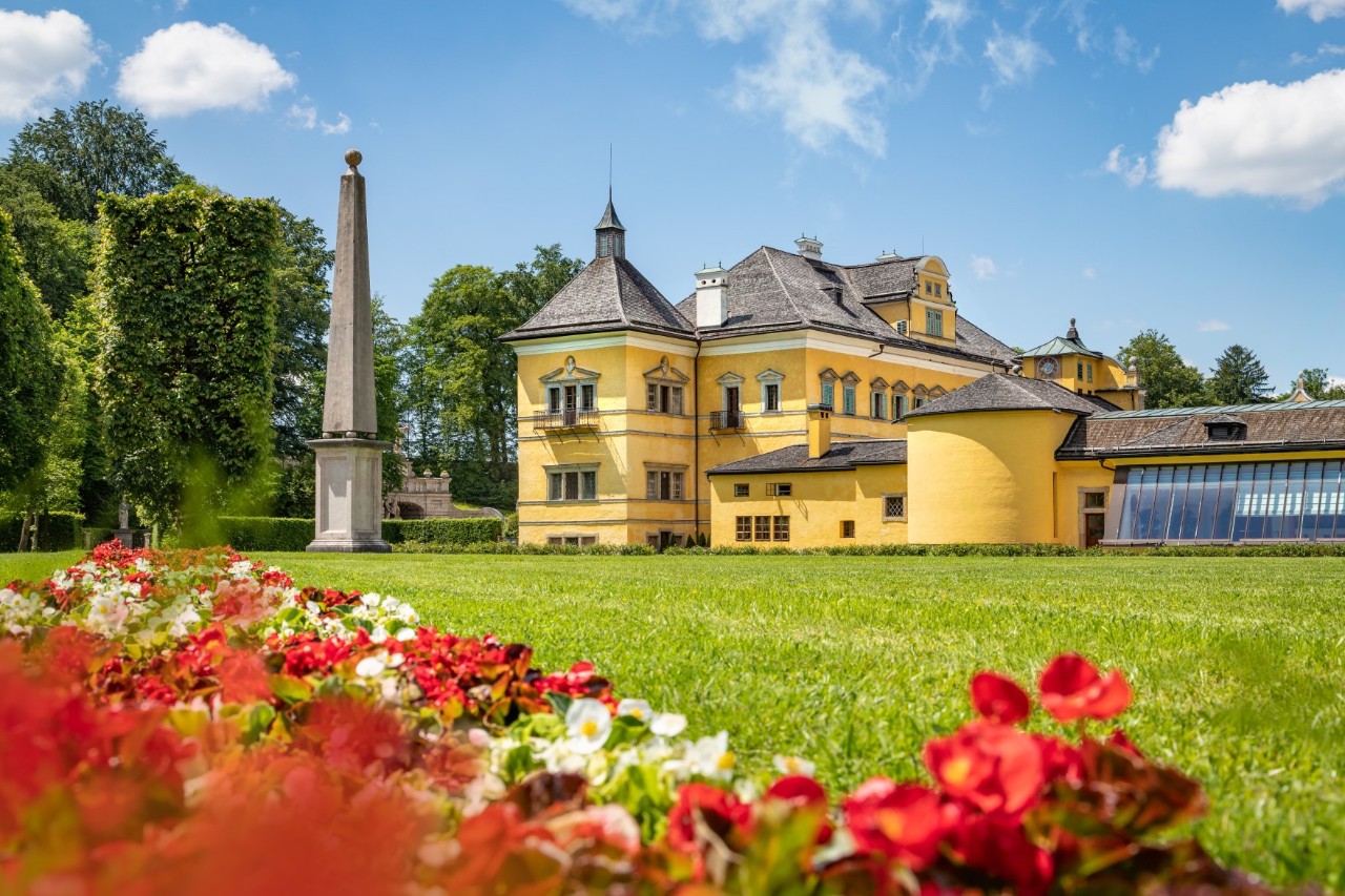 Baroque palace with a yellow front and grey roof in the background, a grey obelisk on the left with several green trees behind it. There is a green lawn in front of the palace and a red and white flowerbed at the front left of the picture.