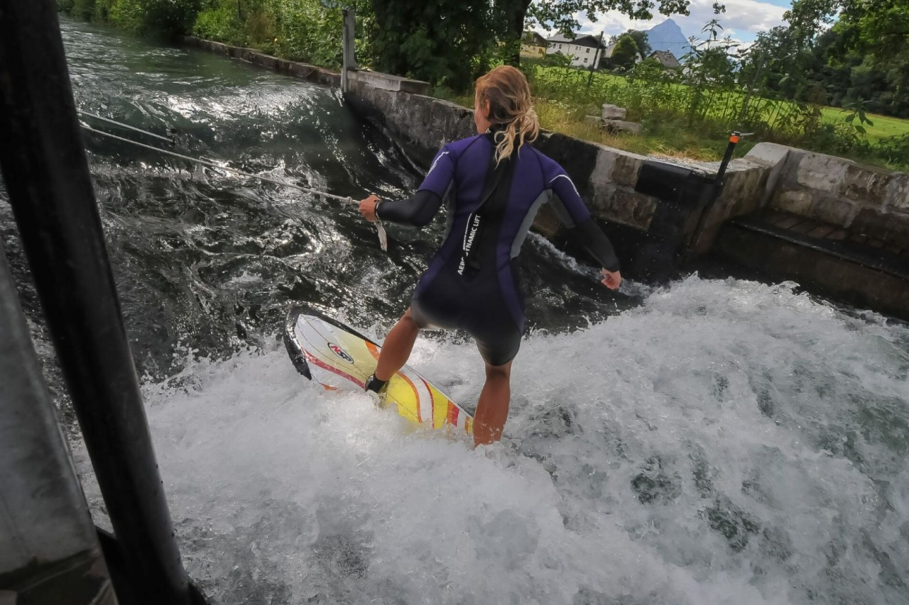 A short, black neoprene suit standing on a surfboard, holding a rope in her left hand, in the middle of a canal in which artificial waves are created.