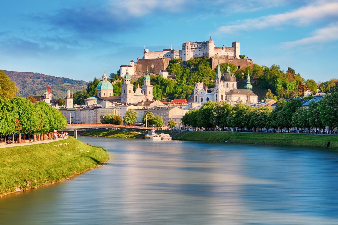 A large castle complex consisting of several buildings with towers and fortress walls and numerous trees in between. A river flows along the valley in the foreground. There is a tree-lined promenade to the left and right of the shore.
