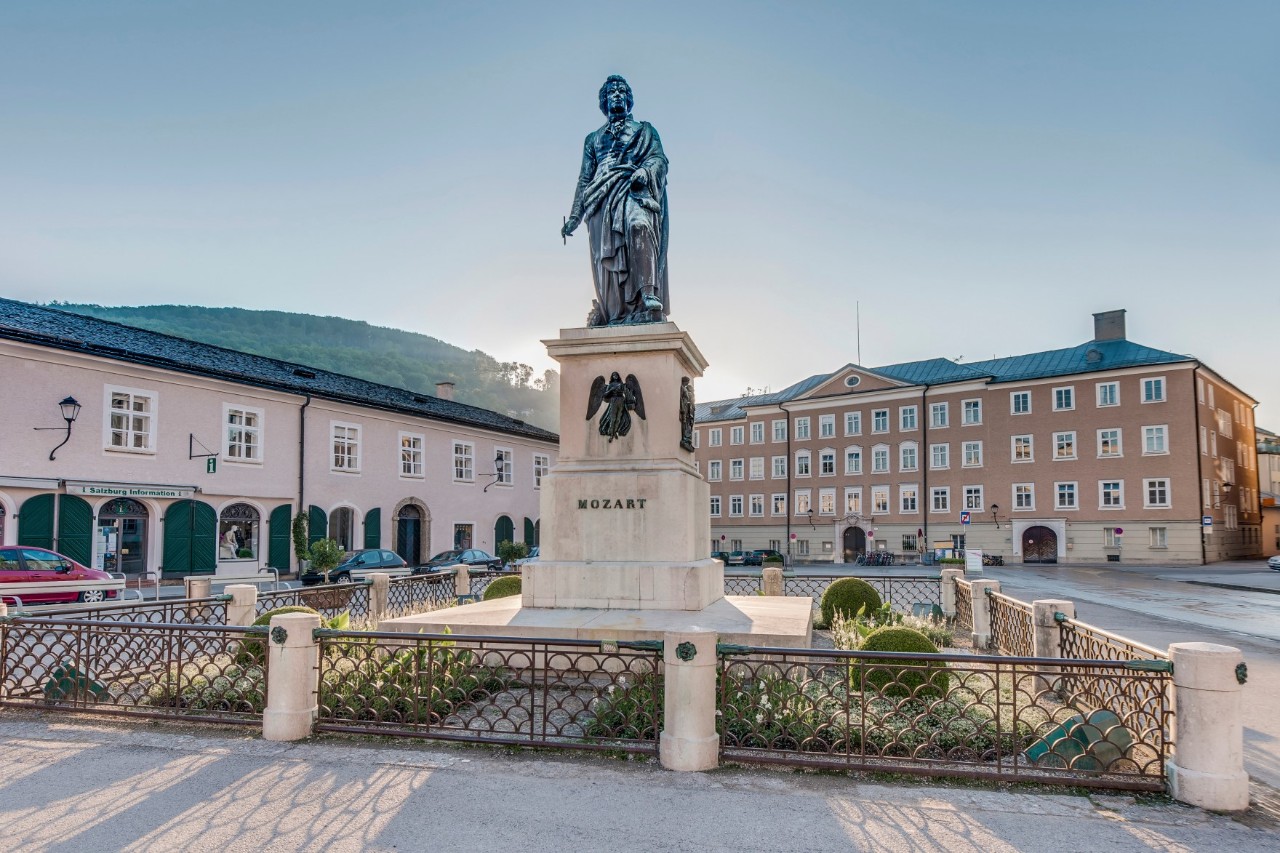 Bronze statue on a sand-coloured pedestal with the inscription “Mozart” surrounded by a small fence laid out as a square, standing on an empty square. Two light-coloured buildings can be seen in the background and there are mountains on the horizon.