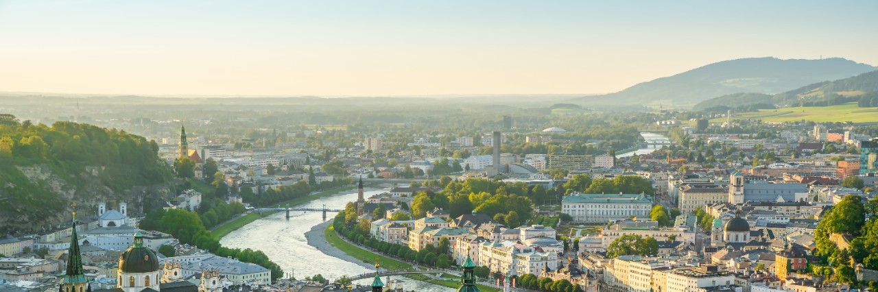 View of the city of Salzburg from above. In the foreground is a large church complex with three towers with domes, bordered by many multi-storey, light-coloured buildings. A wide river winds through the centre of the city. Mountains are visible on the horizon.