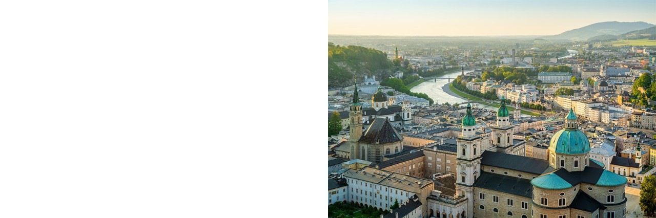 View of the city of Salzburg from above. In the foreground is a large church complex with three towers with domes, bordered by many multi-storey, light-coloured buildings. A wide river winds through the centre of the city. Mountains are visible on the horizon.
