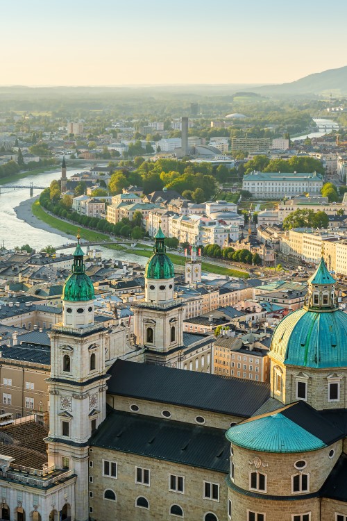 View of the city of Salzburg from above. In the foreground is a large church complex with three towers with domes, bordered by many multi-storey, light-coloured buildings. A wide river winds through the centre of the city. Mountains are visible on the horizon.