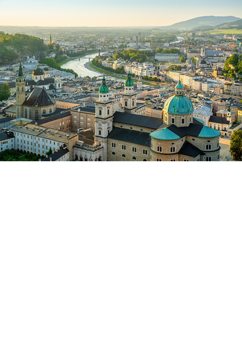 View of the city of Salzburg from above. In the foreground is a large church complex with three towers with domes, bordered by many multi-storey, light-coloured buildings. A wide river winds through the centre of the city. Mountains are visible on the horizon.
