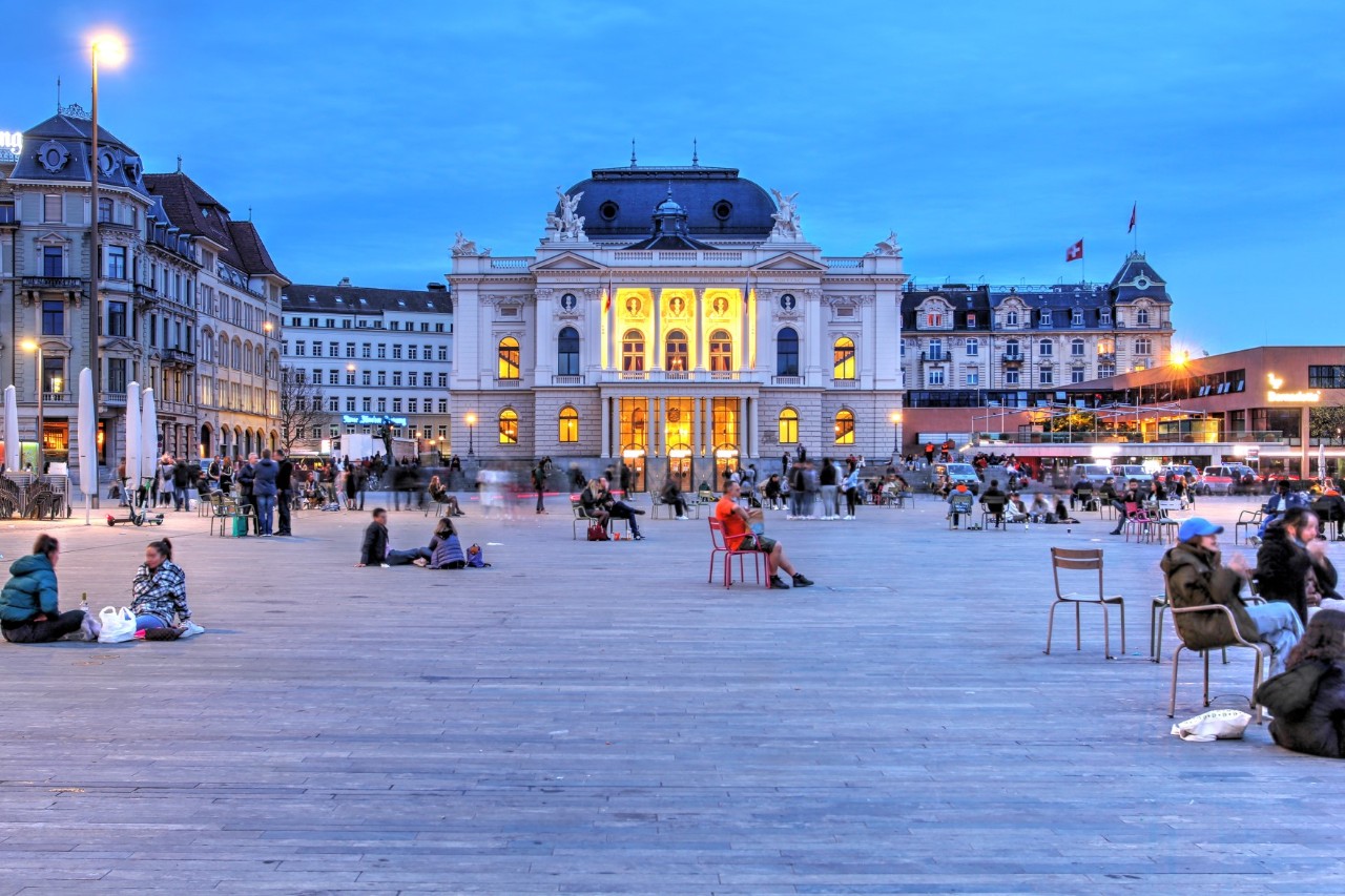 View of a distinctive, bright building, illuminated yellow in the centre and surrounded by other buildings. There is a large square in front of it with people sitting on the ground or on chairs scattered or in groups. It is early dusk.