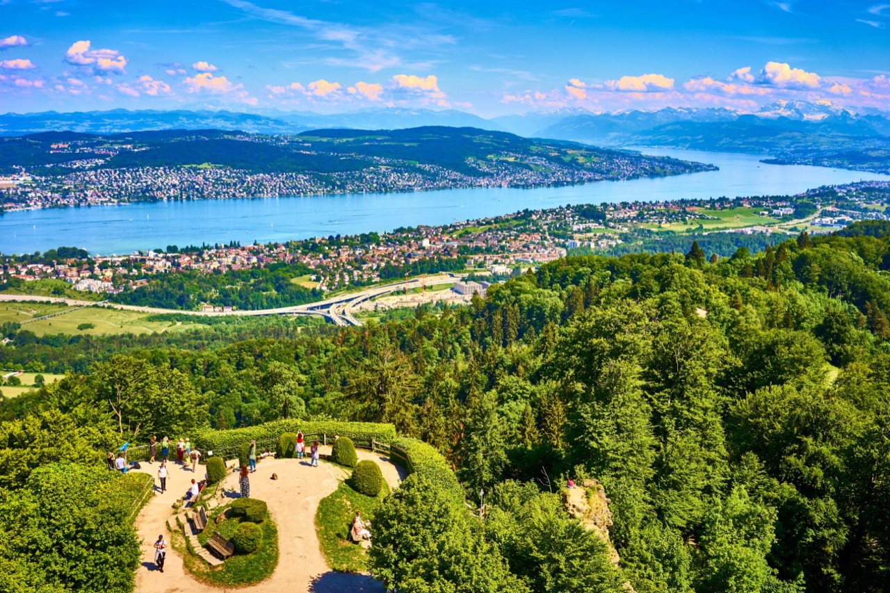 View from above over a forest and a district with lots of flat buildings by a long lake. In the foreground there is a higher viewpoint with some people and a mountain panorama in the background on the other side of the lake.