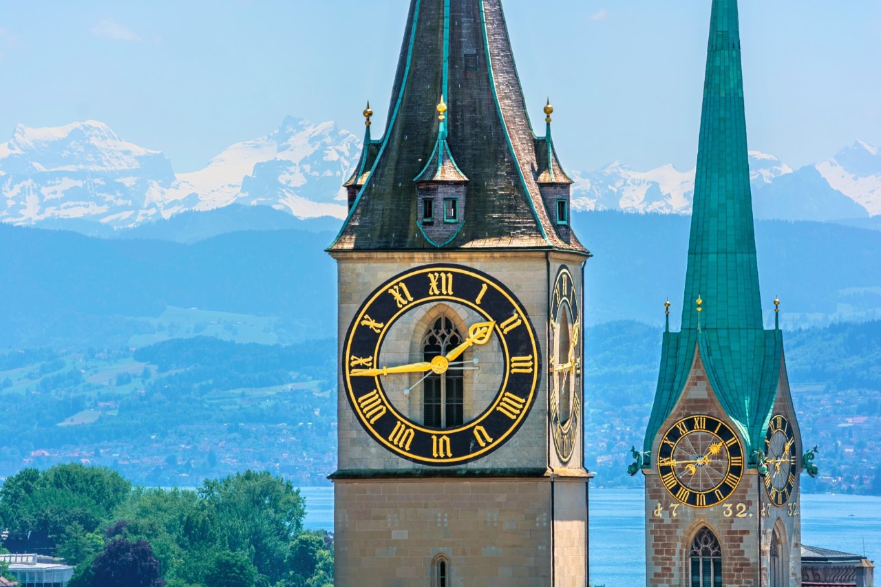 View of two pointed church towers, each with two gilded clock faces with hands. In the background is a lake with an expansive shoreline and a snow-covered mountain panorama behind it.