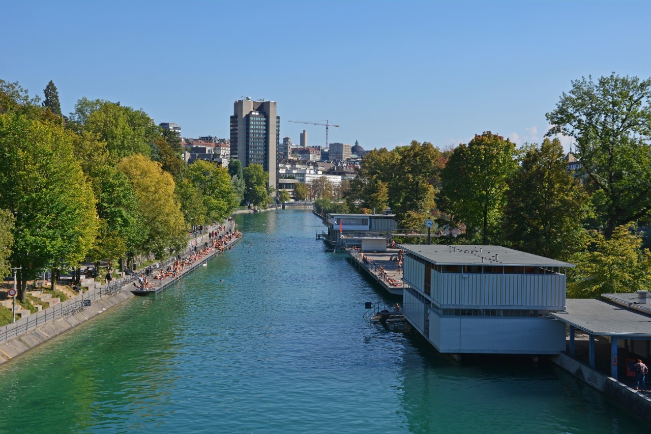 A blue-green river, on the banks of which there are trees and an outdoor swimming pool with a jetty on the right of the picture, and a promenade on the left of the picture. A tower block, several smaller buildings and a crane can be seen at the back of the picture.