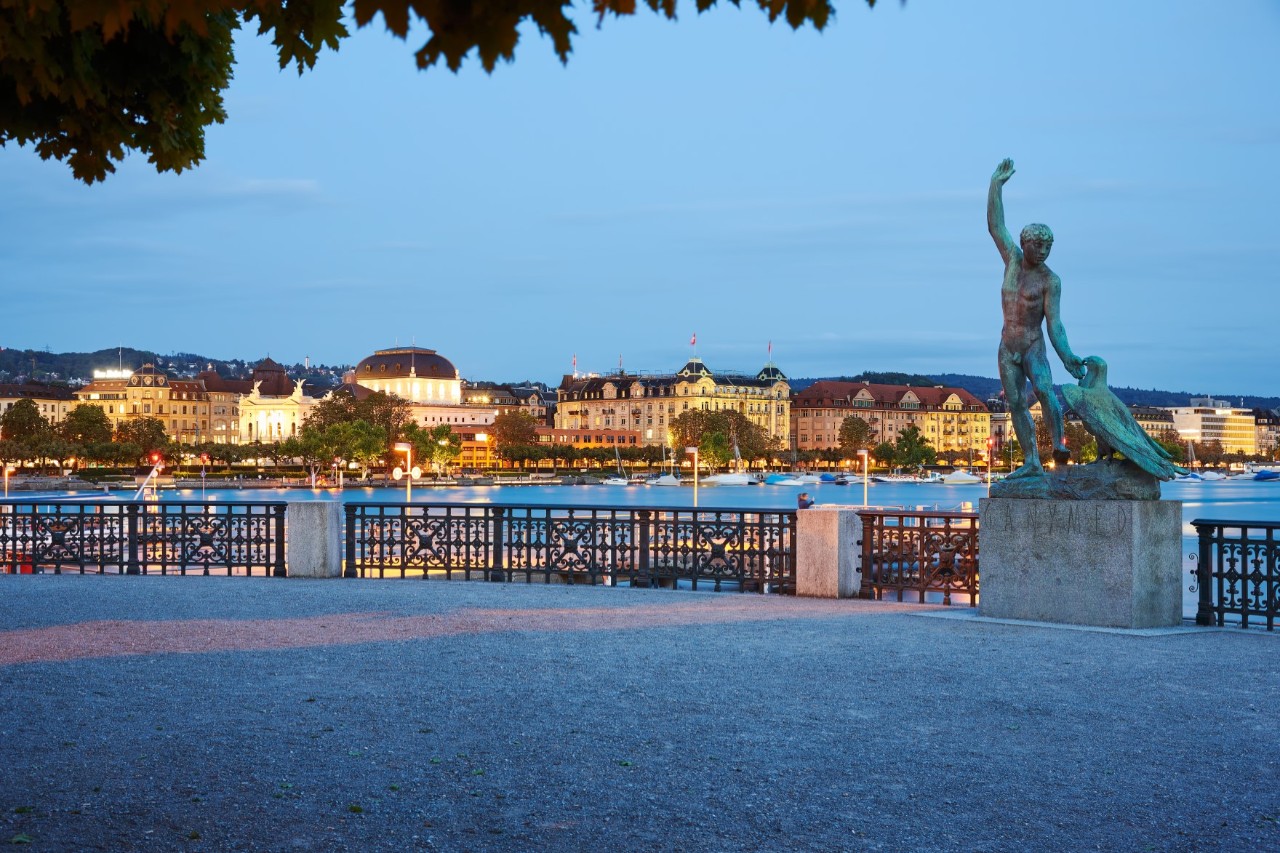 A deserted terrace from which there is a view of a lake and its illuminated shore with buildings on the other side at dusk. A green sculpture of a boy with an eagle stands on a stone at the right of the terrace.