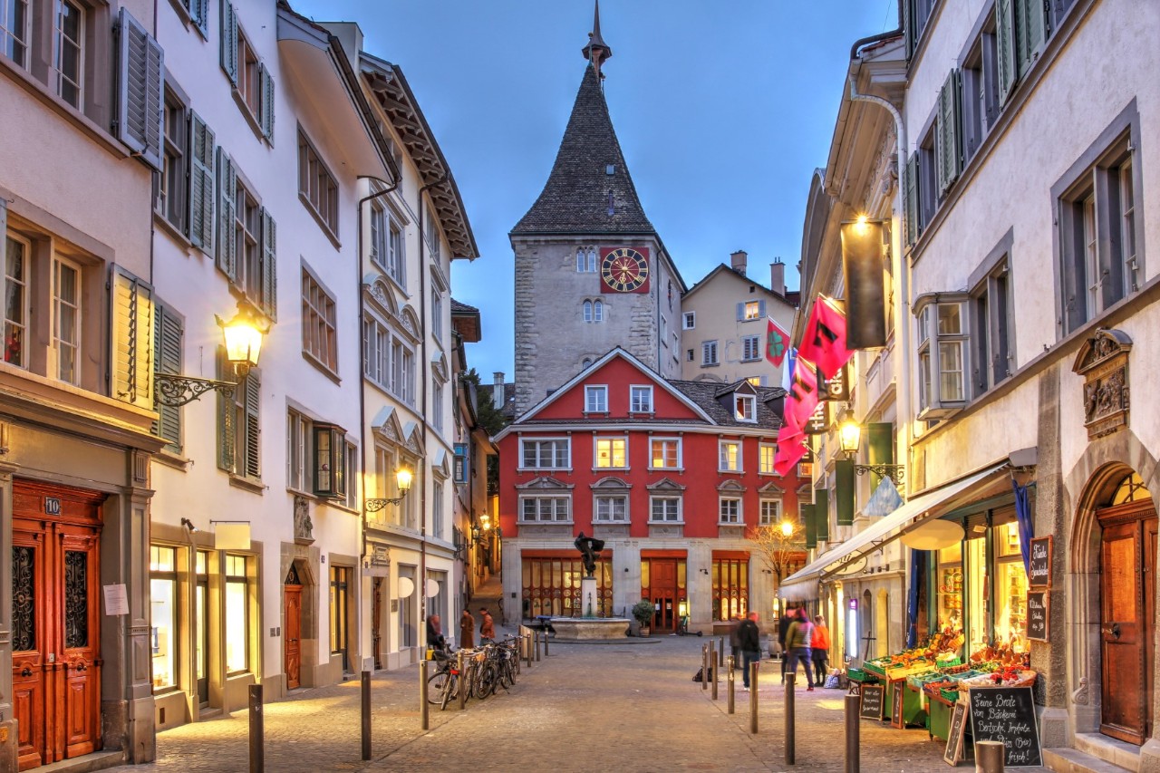 View of an almost deserted old town alleyway, with four-storey buildings with light-coloured fronts on both sides. Straight ahead is a red building and behind it a church tower with a clock face on its front. A small shop with a display of fruit and vegetables can be seen on the right of the picture. Lanterns light the street.