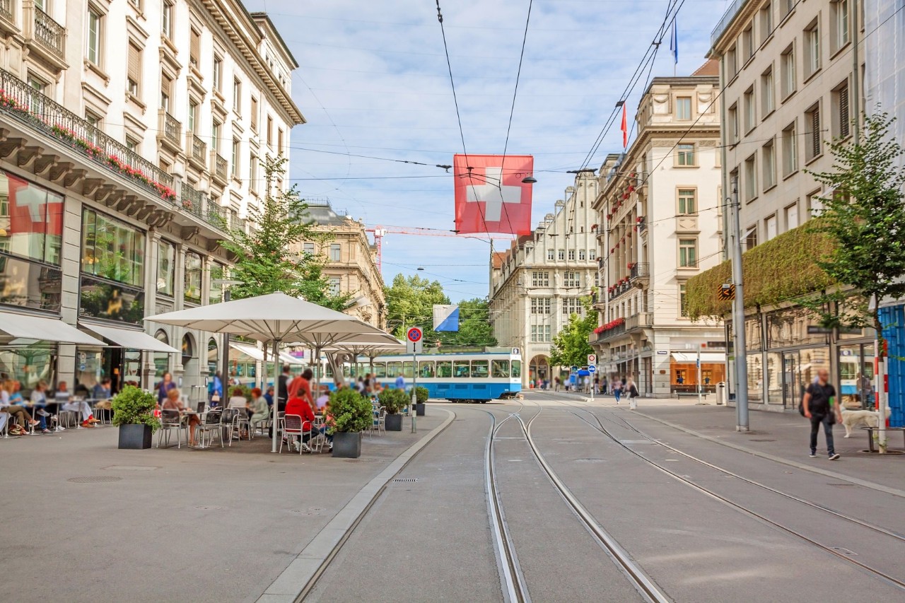 A street with wide footpaths on which few people are walking. In the centre of the picture are tram tracks upon which a blue and white  tram is travelling. A Swiss flag with a white cross on a red background hangs above the tracks. A pavement café with white parasols can be seen on the left.