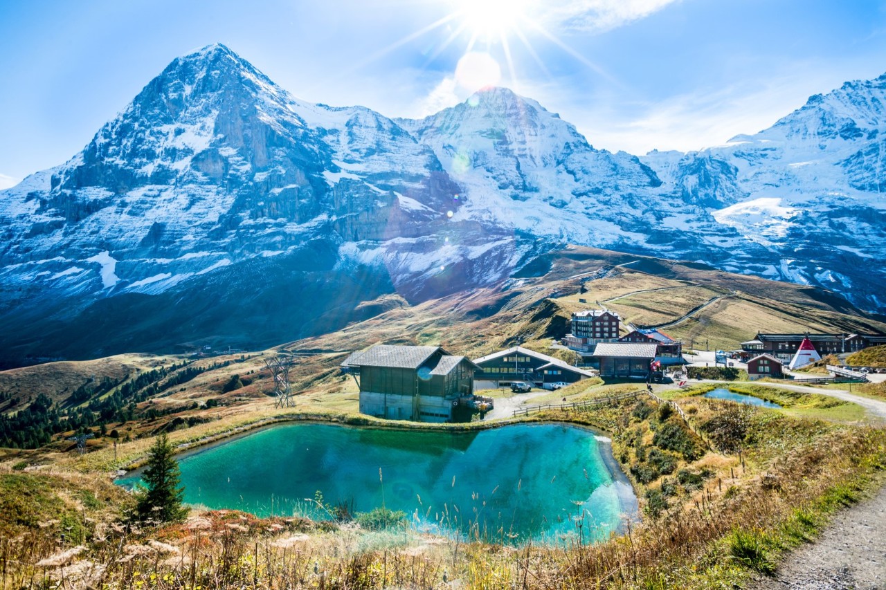 A snow-covered high mountain panorama, behind which the sun is shining. A small turquoise lake can be seen in the foreground surrounded by mountain huts and alpine meadows. To the right is a hiking trail winding its way down into the valley.
