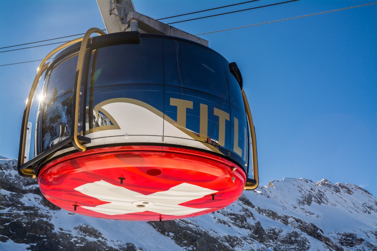 View of a round, glass cable car gondola. The symbol of the Swiss flag with a white cross on a red background can be seen on the underside. The gondola is travelling along snow-covered mountains. The sky is bright blue.