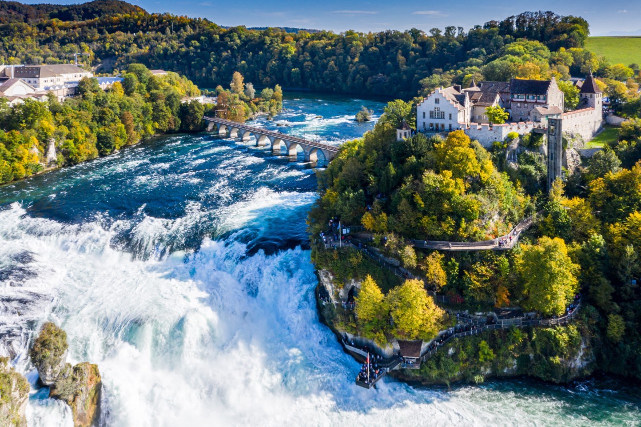 Aerial view of a roaring waterfall, on the right side of which is a castle on a tree-covered hill and a path winding its way to a viewing platform. Above the waterfall is a stone bridge with arches over the river. There is a forest in the background.