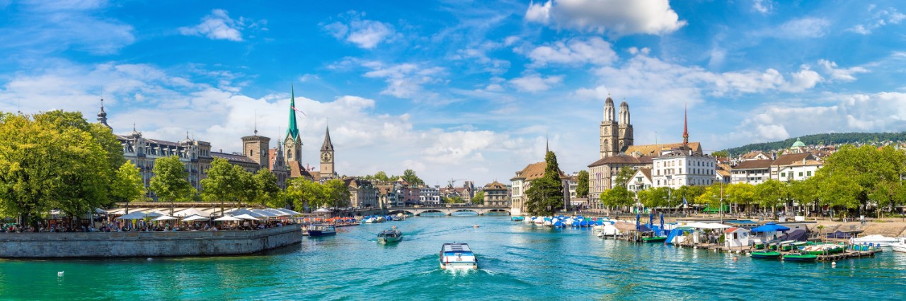 View of a green-blue river running from a lake through the centre of a city. A few small boats are travelling on the river. Trees, building and church towers can be seen on both river banks. There is a lively beer garden with parasols on the left bank at the front. Boats and landings can be seen on the front right bank.