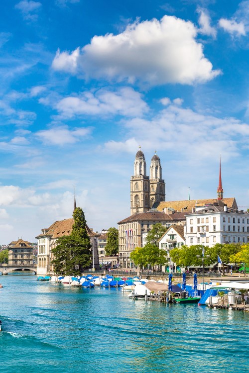 View of a green-blue river running from a lake through the centre of a city. A few small boats are travelling on the river. Trees, building and church towers can be seen on both river banks. There is a lively beer garden with parasols on the left bank at the front. Boats and landings can be seen on the front right bank.