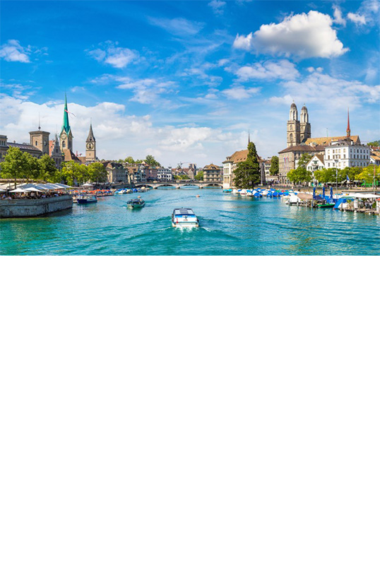 View of a green-blue river running from a lake through the centre of a city. A few small boats are travelling on the river. Trees, building and church towers can be seen on both river banks. There is a lively beer garden with parasols on the left bank at the front. Boats and landings can be seen on the front right bank.