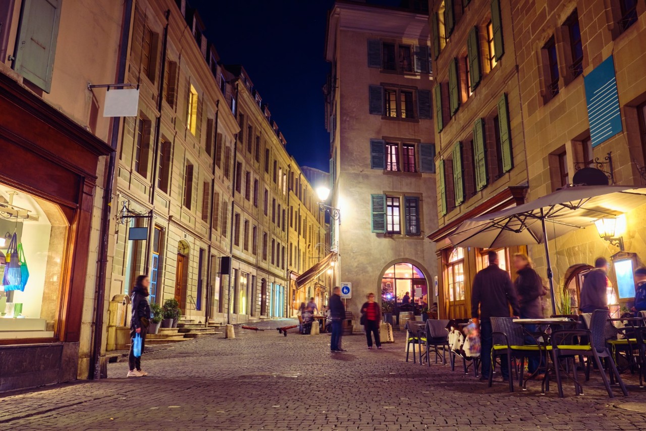 An illuminated, car-free street in an old town with four-storey, light brown buildings, some of which contain shop windows. A few people are standing in the street. Other people can be seen standing in street cafés on the right of the picture.