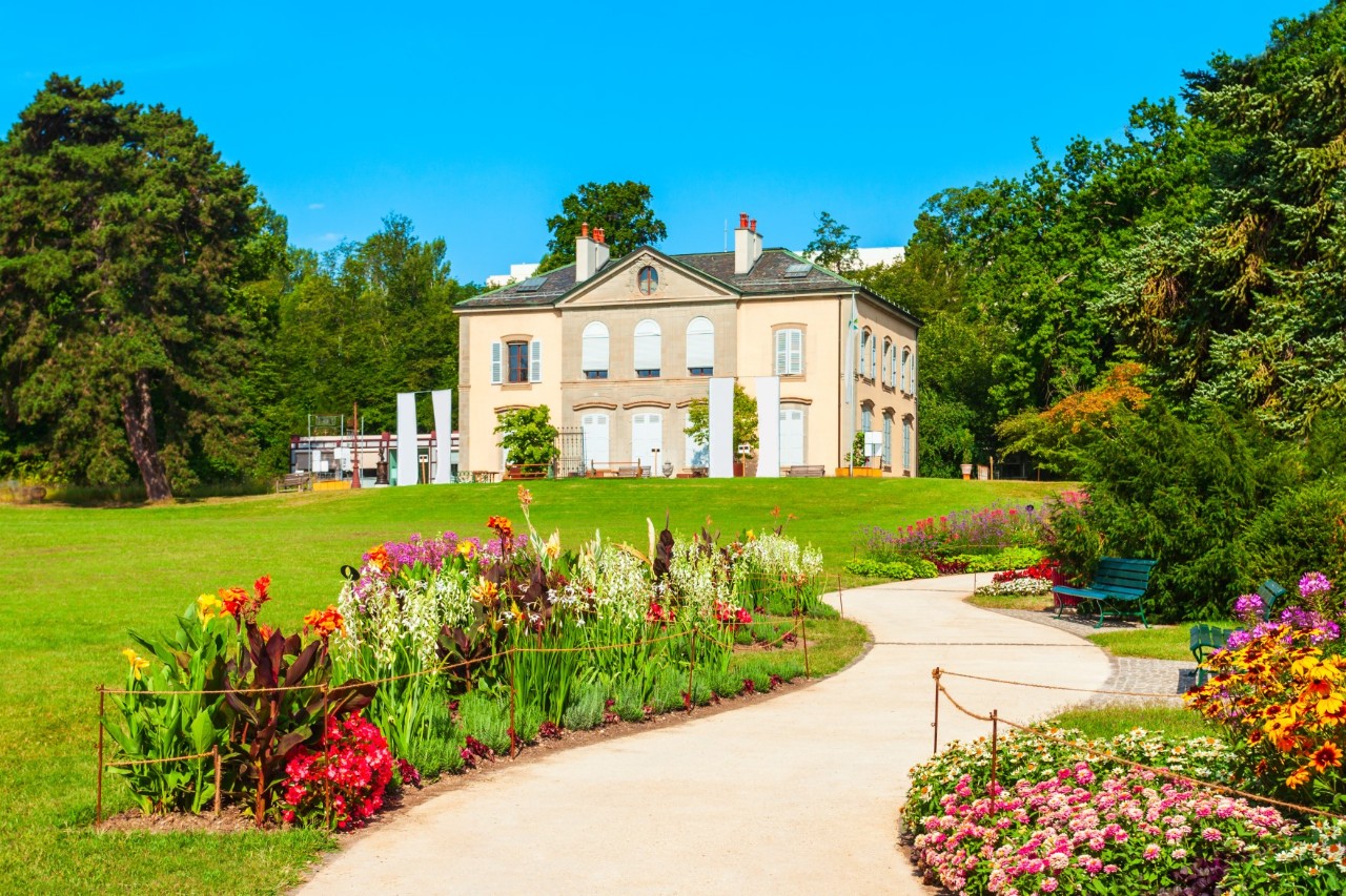 Central view of a white, two-storey building in a well-maintained park with trees to the left and right of the building, a lawn in front and colourful flowerbeds. A light-coloured path with benches along the side meanders towards the building.