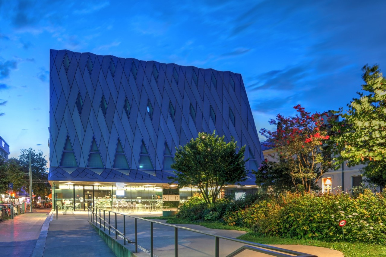 View of a modern building with a towering pointed roof, made of glass and metal; it stands out thanks to the elegant aesthetics of its striking geometric pattern. The picture was taken at dusk during the blue hour. A deserted footpath can be seen in front of the building, with trees and shrubs planted along the edge.