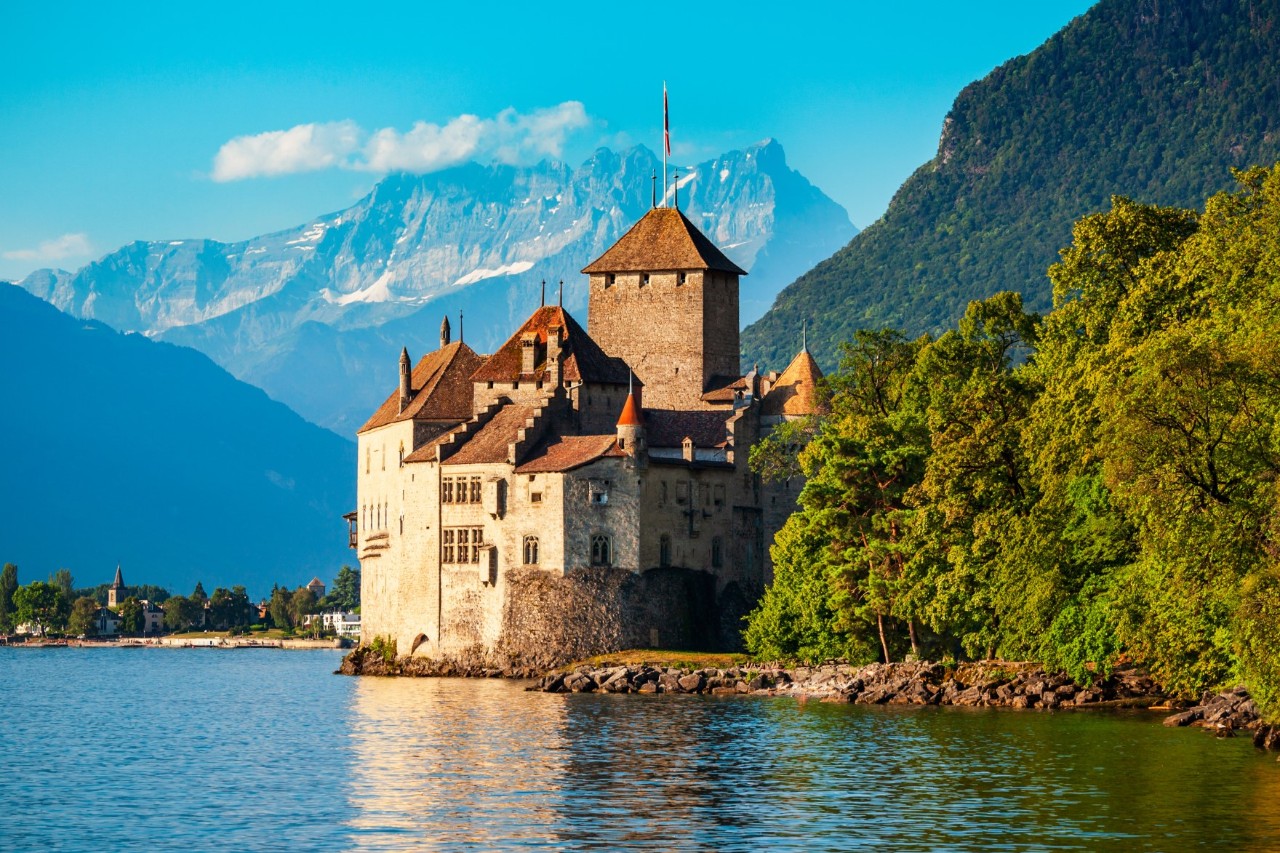 View of an old castle standing directly on the shore of a lake surrounded by high mountains in the background and green, dense forest to the right of the picture. Glaciers can be seen in the mountains.