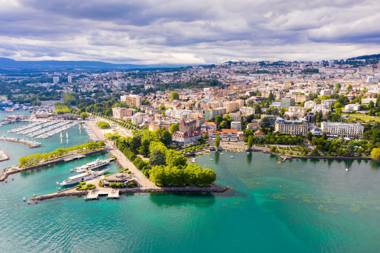 A sprawling city with multi-storey buildings and trees in between. In the foreground of the picture is a green-blue lake with green overgrown headlands and a small harbour.