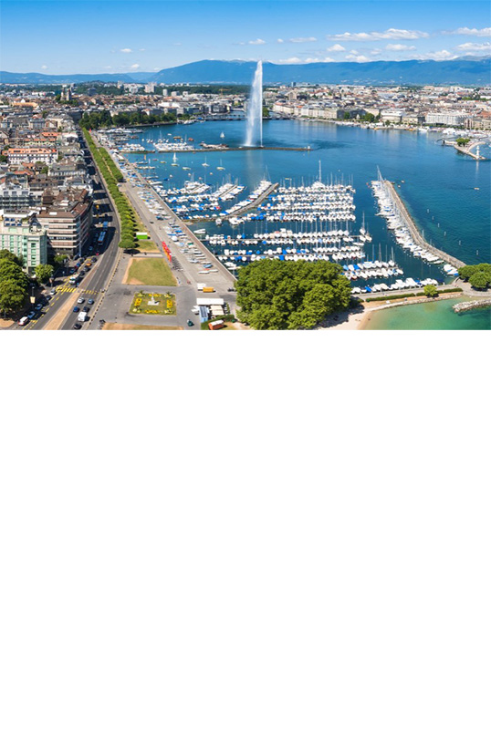 Aerial view of a city located directly on a green-blue lake, in which there is a harbour with many small boats and a water fountain. Flat mountains can be seen in the background. A road lined with trees runs along the shore.