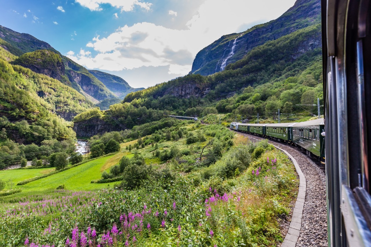 Flåm Railway on winding track between green slopes and rocky terrain.