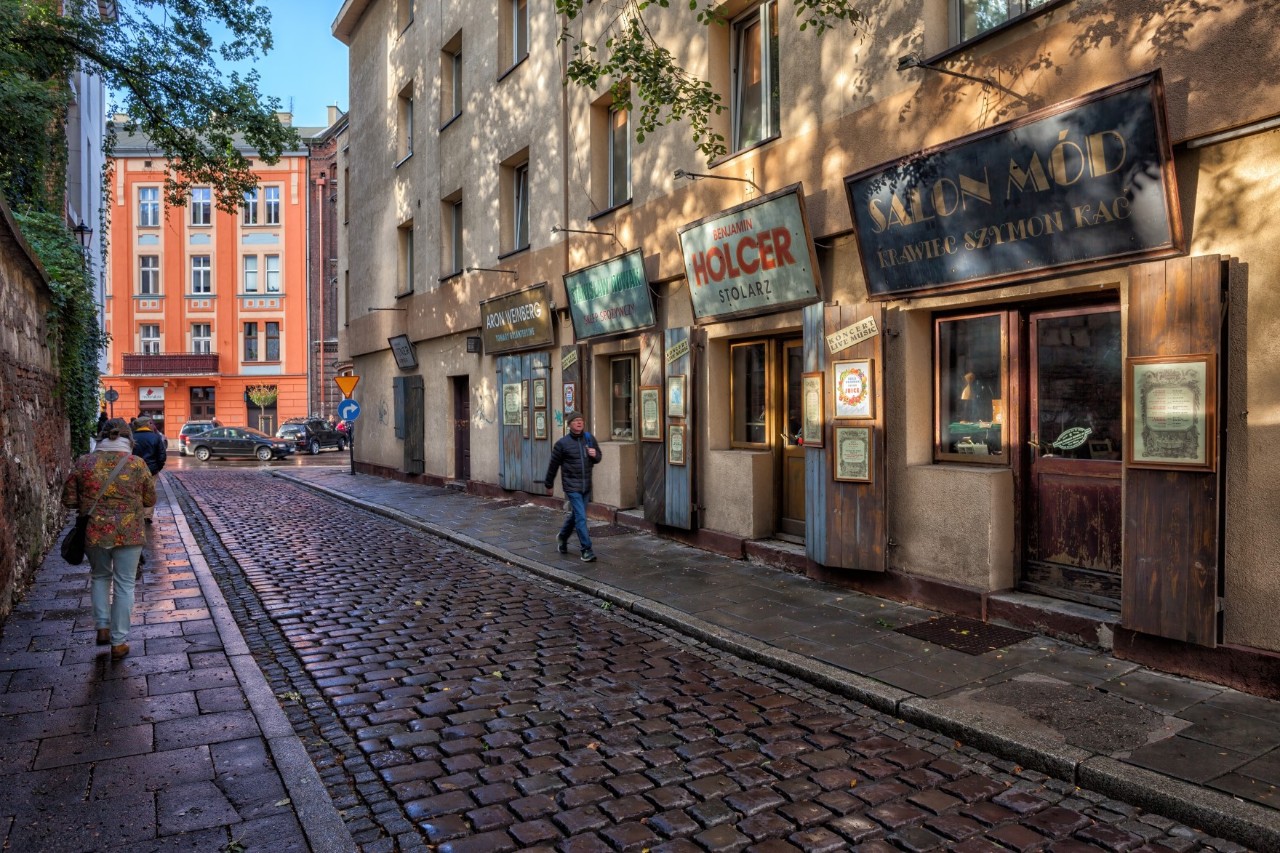A cobbled street, shops can be seen on the right-hand side of the street, a wall can be seen on the left, pedestrians are walking along the street 