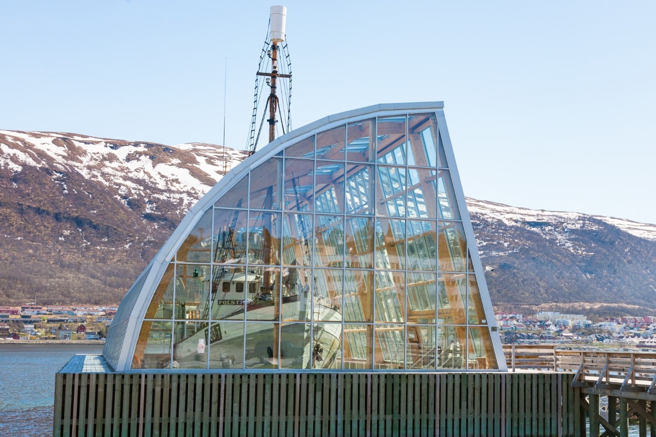 Glass pavilion with reflected boat and snowy mountains in Tromsø.