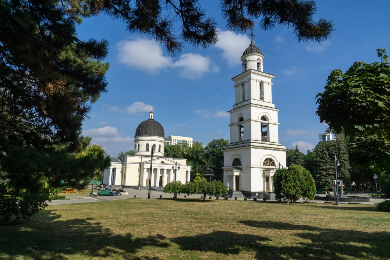 Triumphal arch with clock on a street, white church behind it 