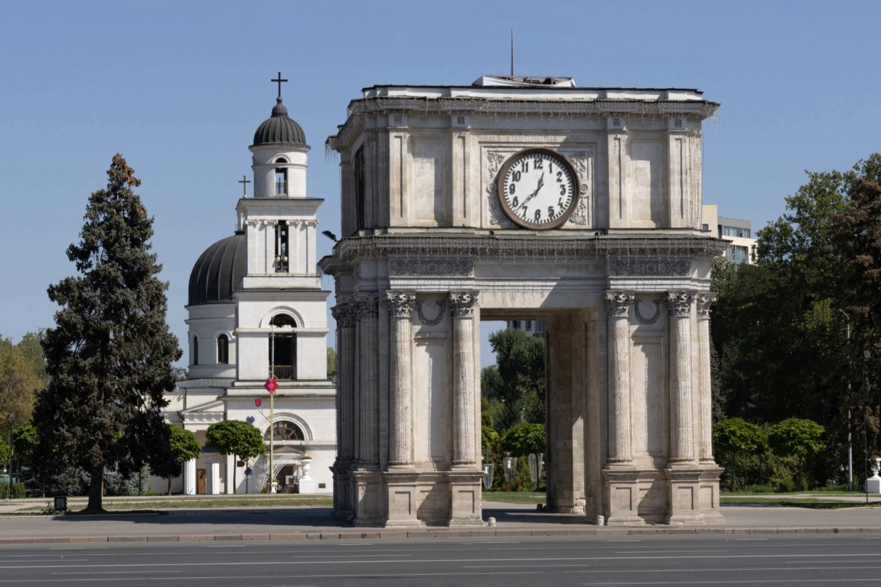 Triumphal arch with clock on a street, white church behind it 